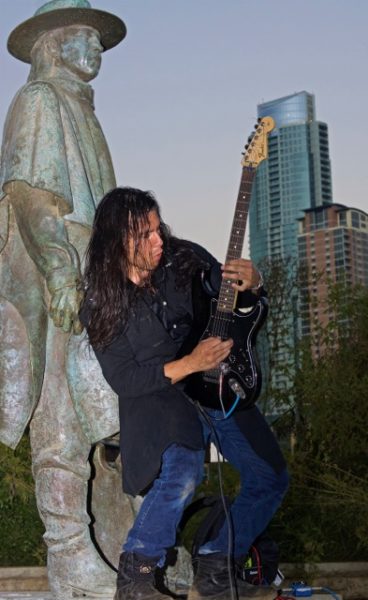 Long haired man playing electric guitar in front of green copper statue