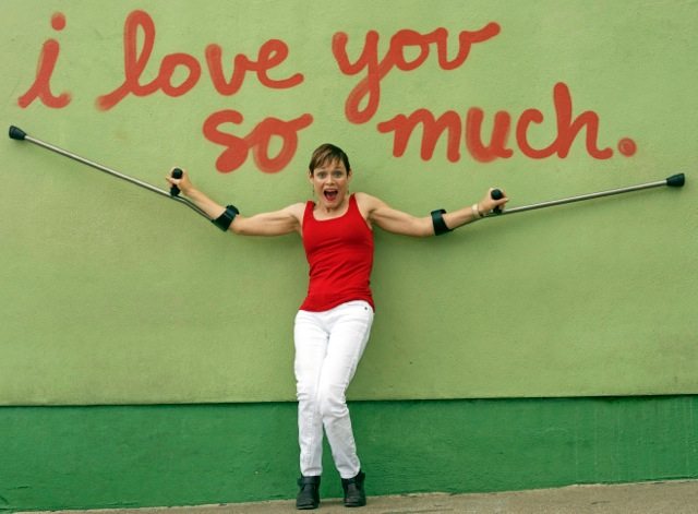 Woman in red shirt holding up arm brace crutches in front of Austin's "i love you so much" spray painted wall